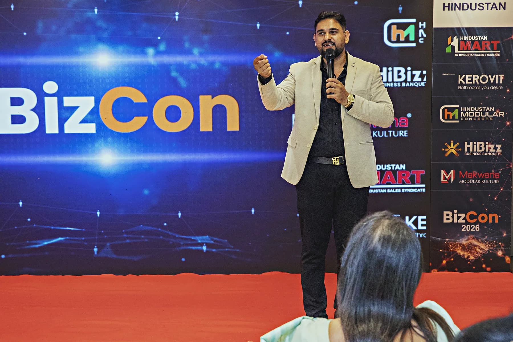 Host speaking on stage against the branded backdrop at the HiBizz launch in Goa